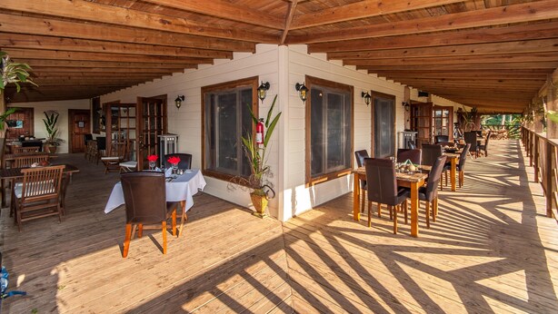 Un espacio exterior para comer con mesas y sillas de madera, ventanas amplias y techo de madera.