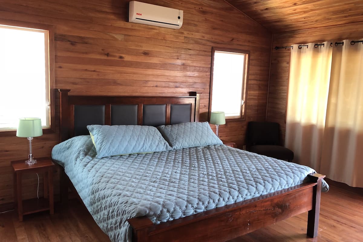 A wooden-paneled bedroom with a bed, two bedside lamps, a window, and an air conditioning unit.