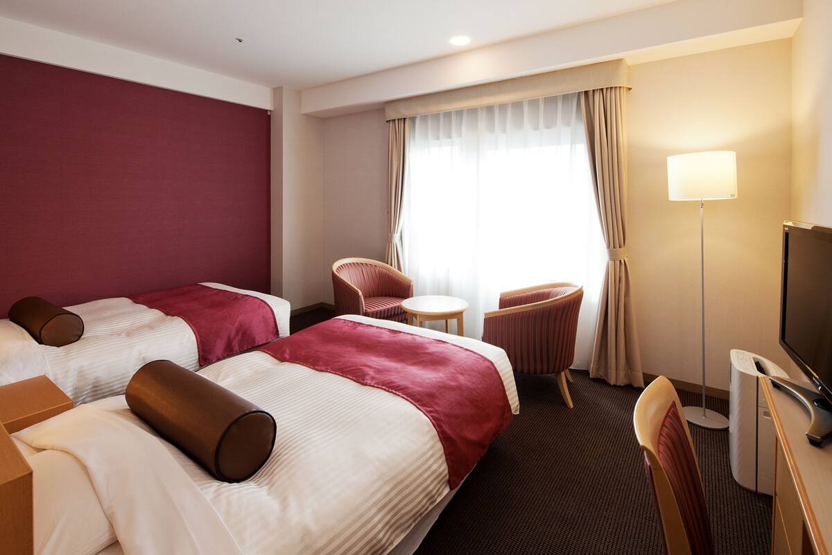 A hotel room with two beds, a chair, a small table, a TV, and a lamp.