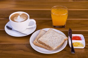 Un desayuno con una taza de café, dos rebanadas de pan, mantequilla, mermelada y un vaso de zumo de naranja sobre una mesa de madera.