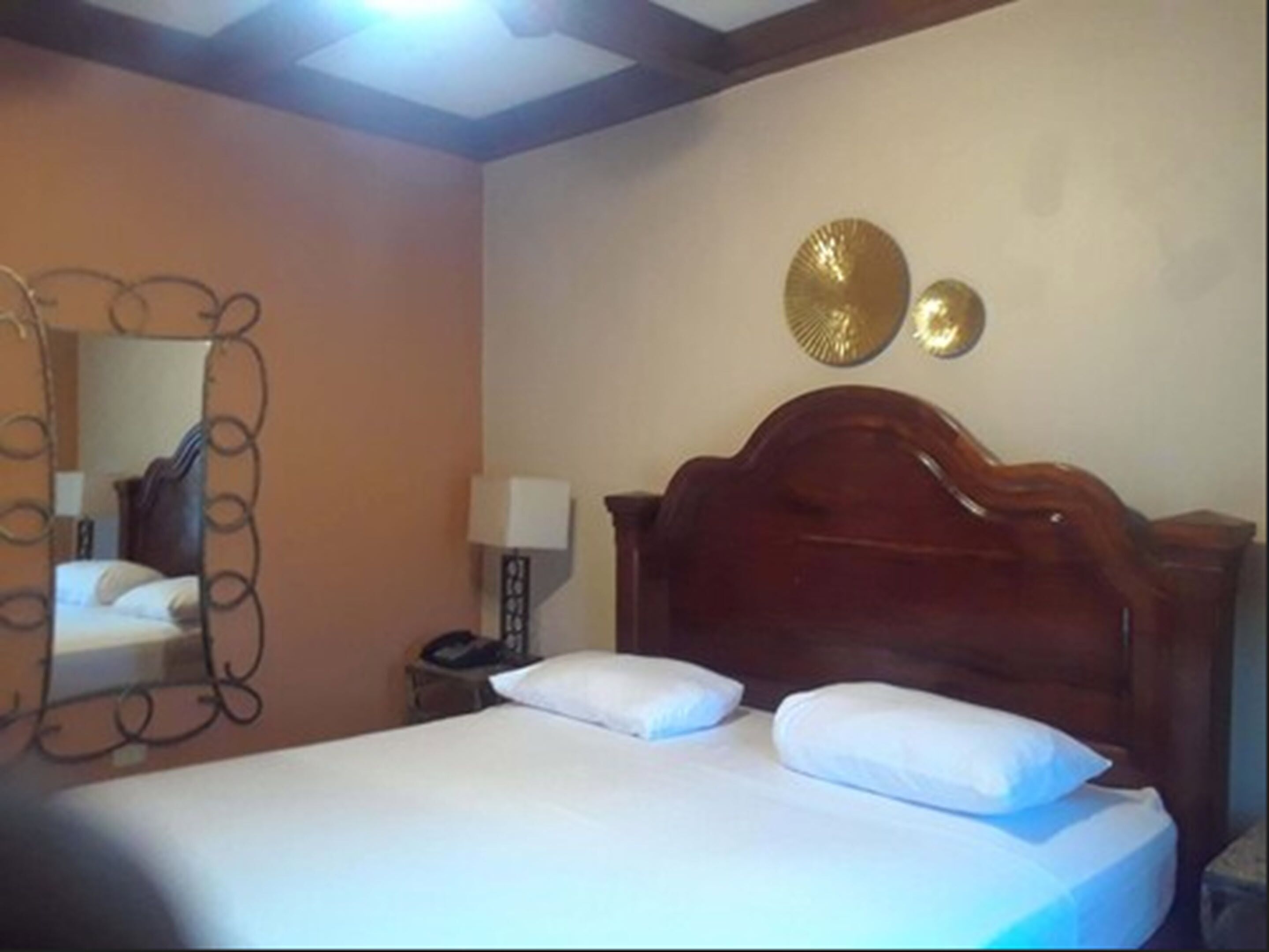 Ein Hotelzimmer mit einem hölzernen Bett, zwei weißen Kissen, einem Nachttisch mit Lampe und einem an der Wand befestigten Spiegel.