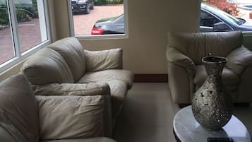 Una sala de estar con un sofá color beige, un jarrón estampado sobre una mesa redonda, y amplias ventanas con vistas a un estacionamiento.