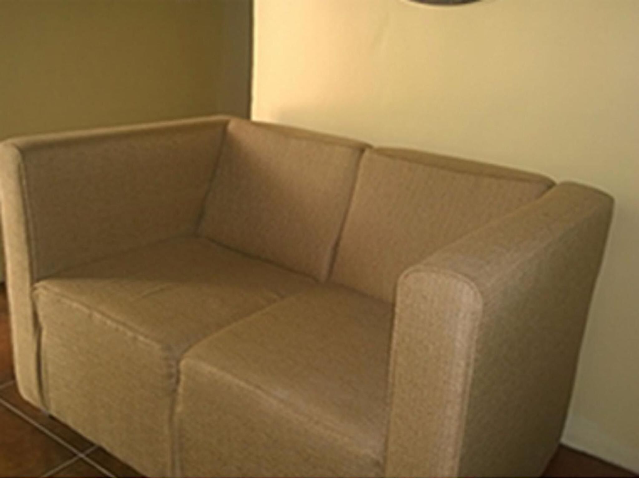 A beige sofa in a corner of a room with plain walls and a tiled floor.