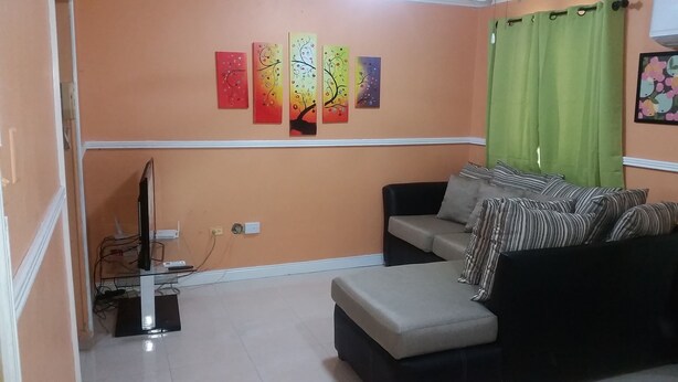 A living room with a sofa, ottoman, and a flat-screen TV. There are four wall-mounted paintings above the sofa.