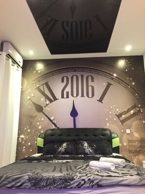 A bedroom with a large clock displaying the year 2016, a tufted headboard, and a bed with patterned bedding.