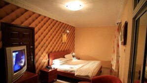 A hotel room with a tufted headboard, a bed with white linens, a television on a wooden stand, a red armchair, and a glass door leading to a bathroom. - Vines Hotel (Abuja)