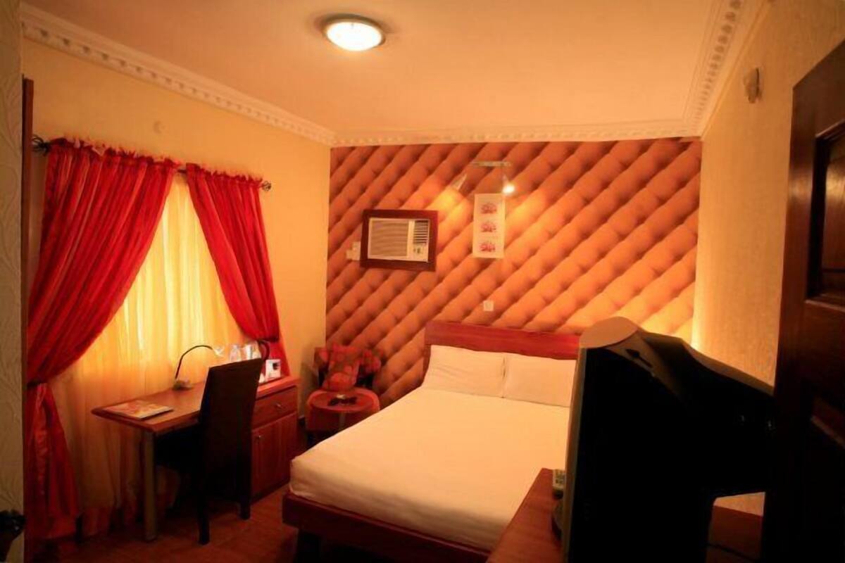 A hotel room with a bed, a desk, and a chair. There is a window with red curtains, a wall-mounted air conditioner, and a framed picture on the wall.