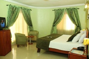 A hotel room with a bed, a chair, a small table, and curtains. - Vines Hotel (Abuja)