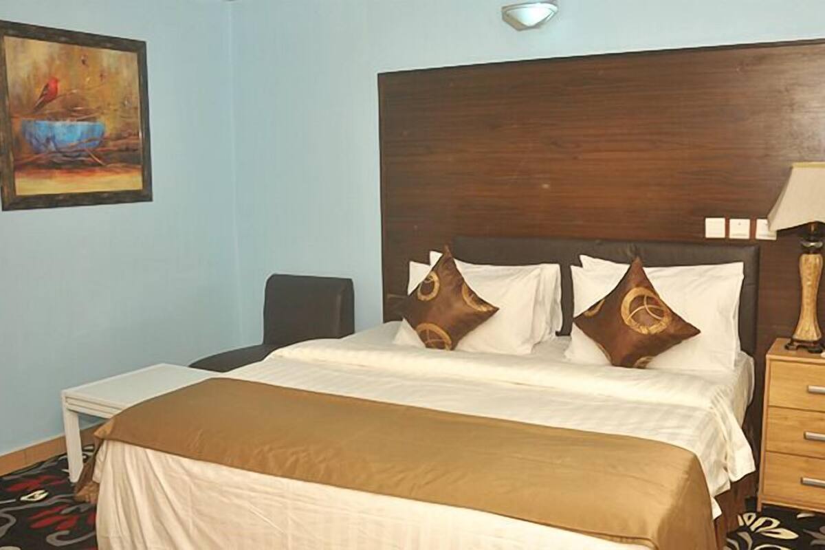 A hotel room with a large bed, a bedside table with a lamp, a small bedside table with a phone, and a painting on the wall.