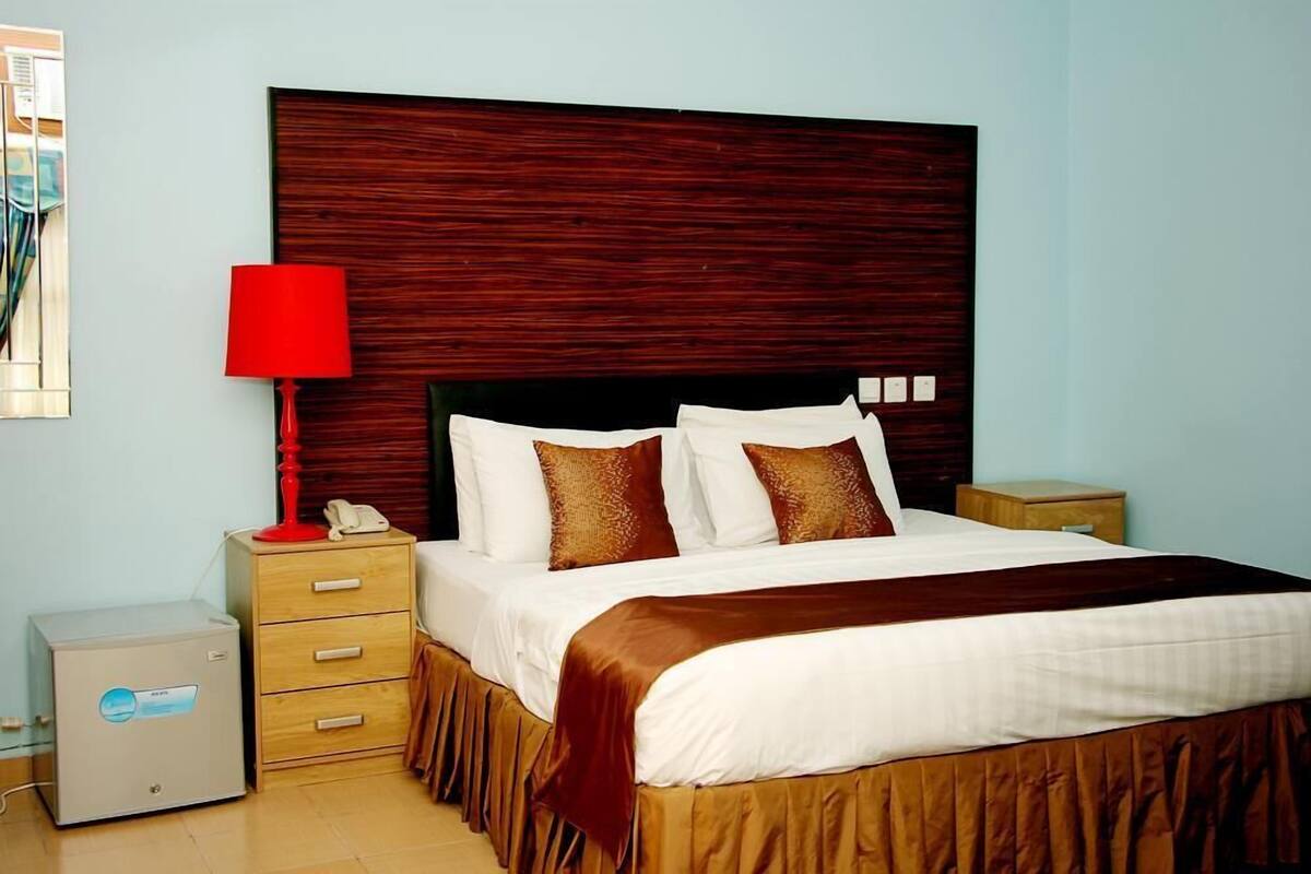 A hotel room with a large bed, a wooden headboard, a red lamp, a bedside table, and a small cabinet.
