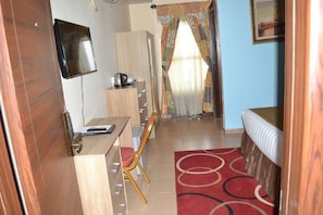 A compact room with a bed, a desk, a chair, a TV, and a window with curtains.