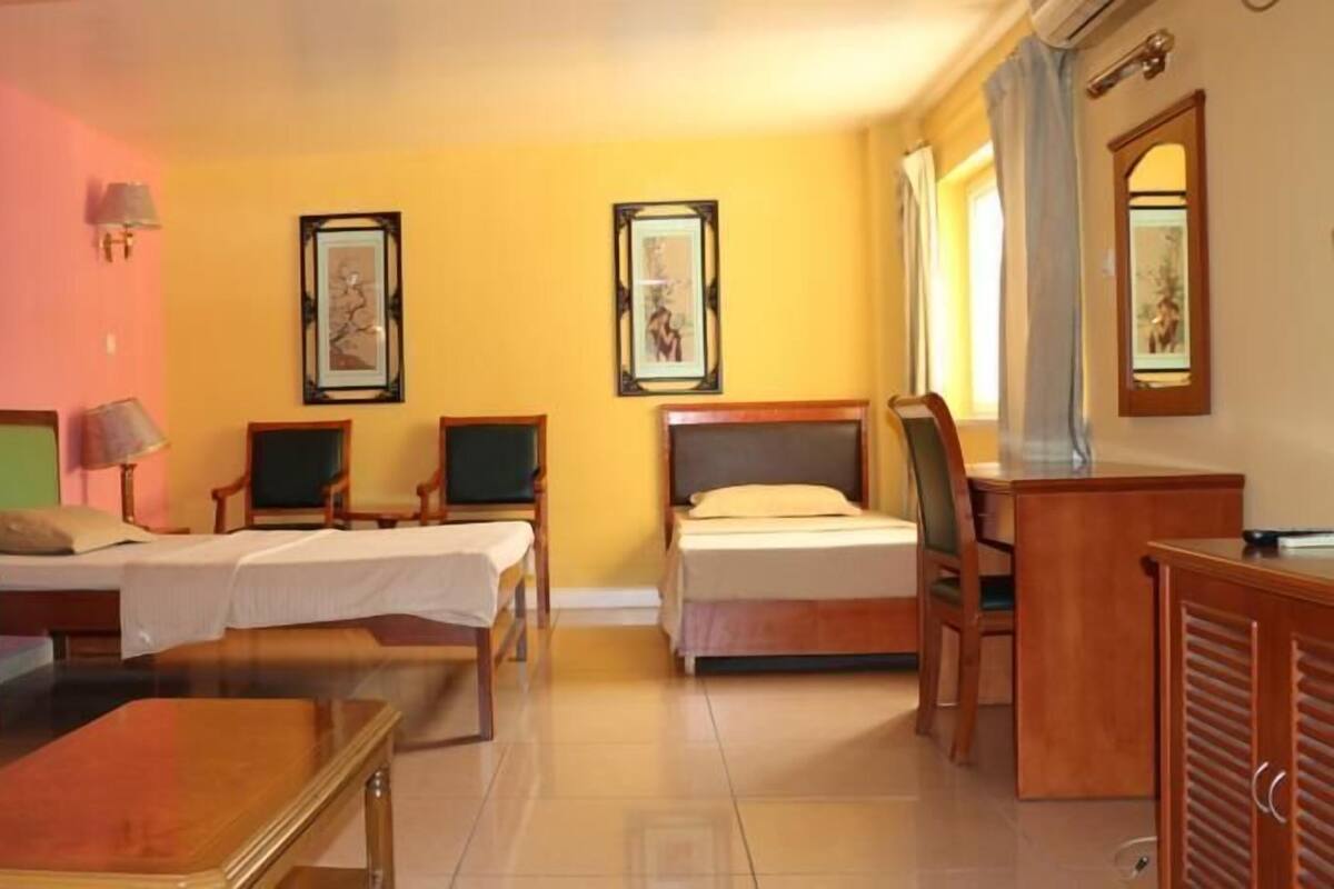 A hotel room with two single beds, a desk, a chair, a small table, and two wall-mounted lamps.