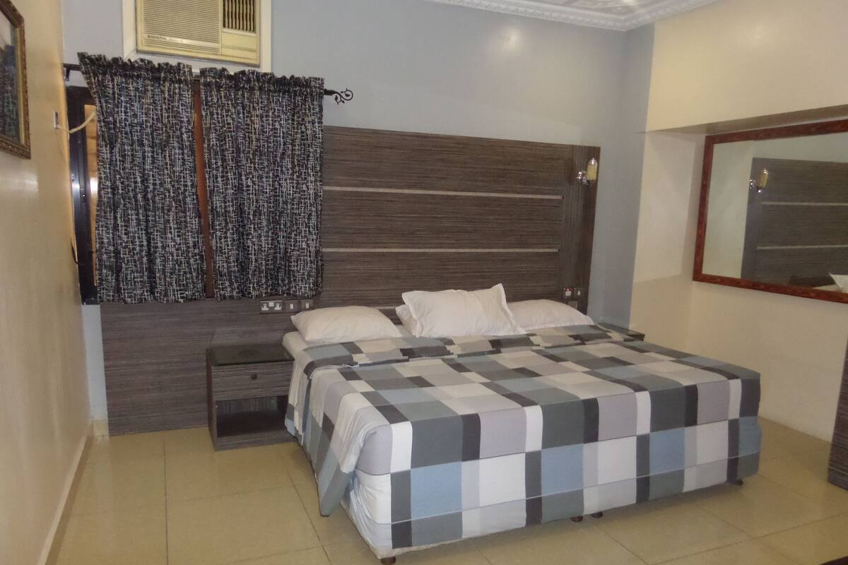 A bedroom with a large bed, a nightstand, a dresser, and a window with patterned curtains.
