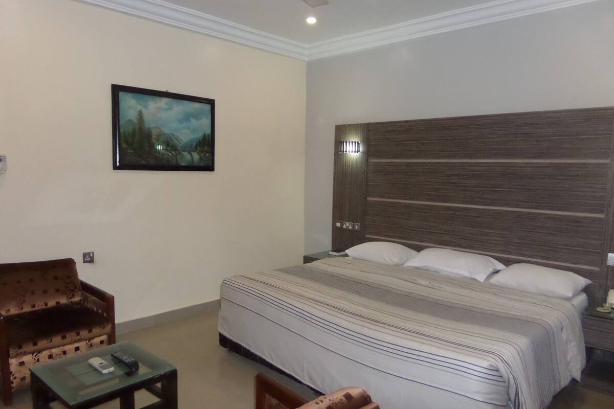 A hotel room with a large bed, a small table, two armchairs, a painting on the wall, and a wooden headboard.