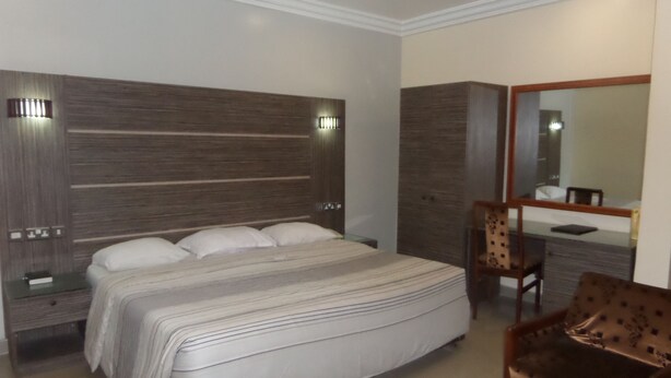 A modern hotel room with a large bed, a wooden headboard, a bedside table, a chair, a small table with remote controls, and a mirror. - Parkview Hotel Abuja (Abuja)
