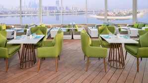 Un comedor moderno con sillas y mesas verdes, amplias ventanas con vista a la ciudad y suelo de madera tipo terraza.