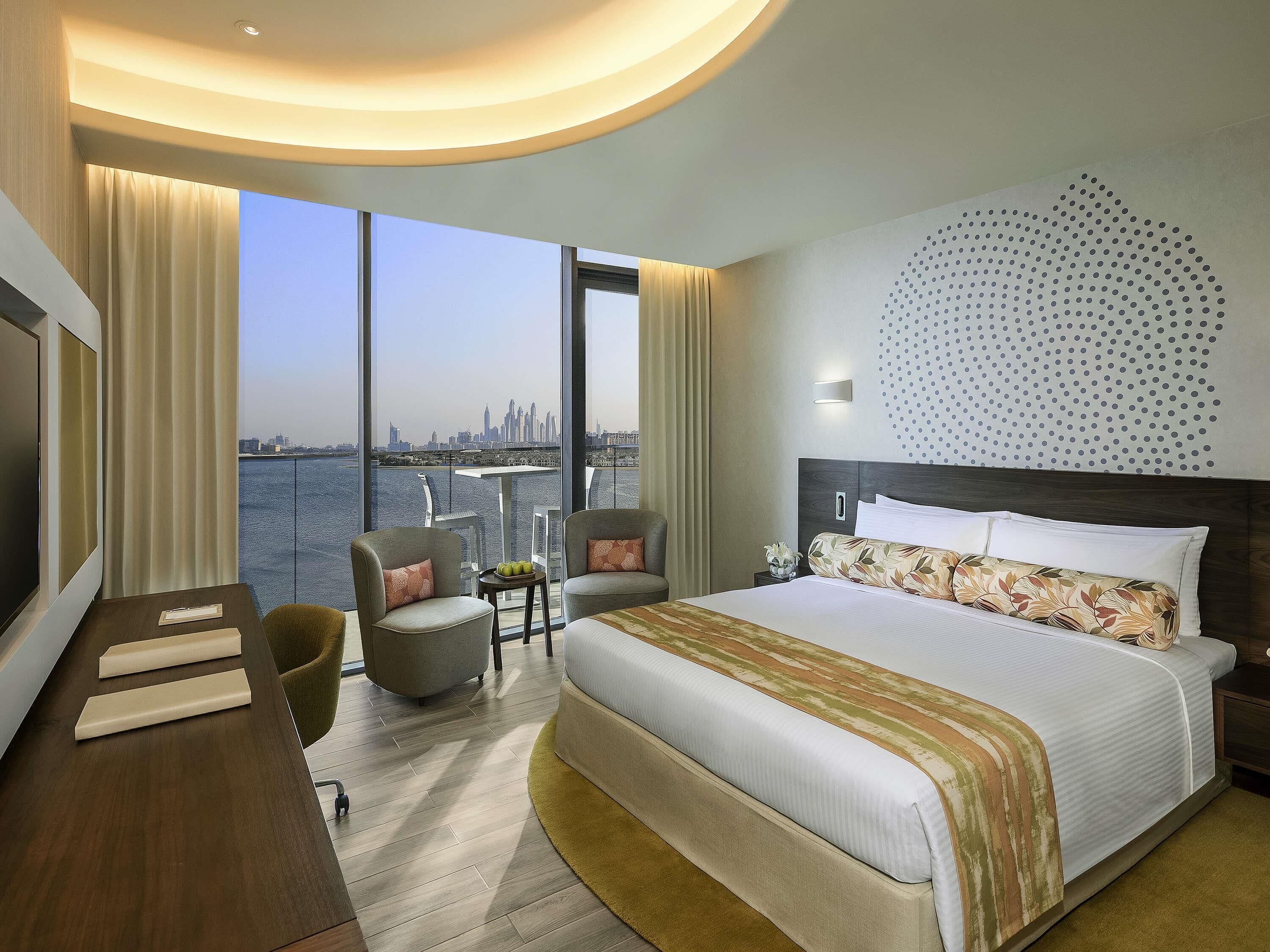 Une chambre d’hôtel avec un grand lit, deux fauteuils, un bureau et une vue sur le panorama urbain.