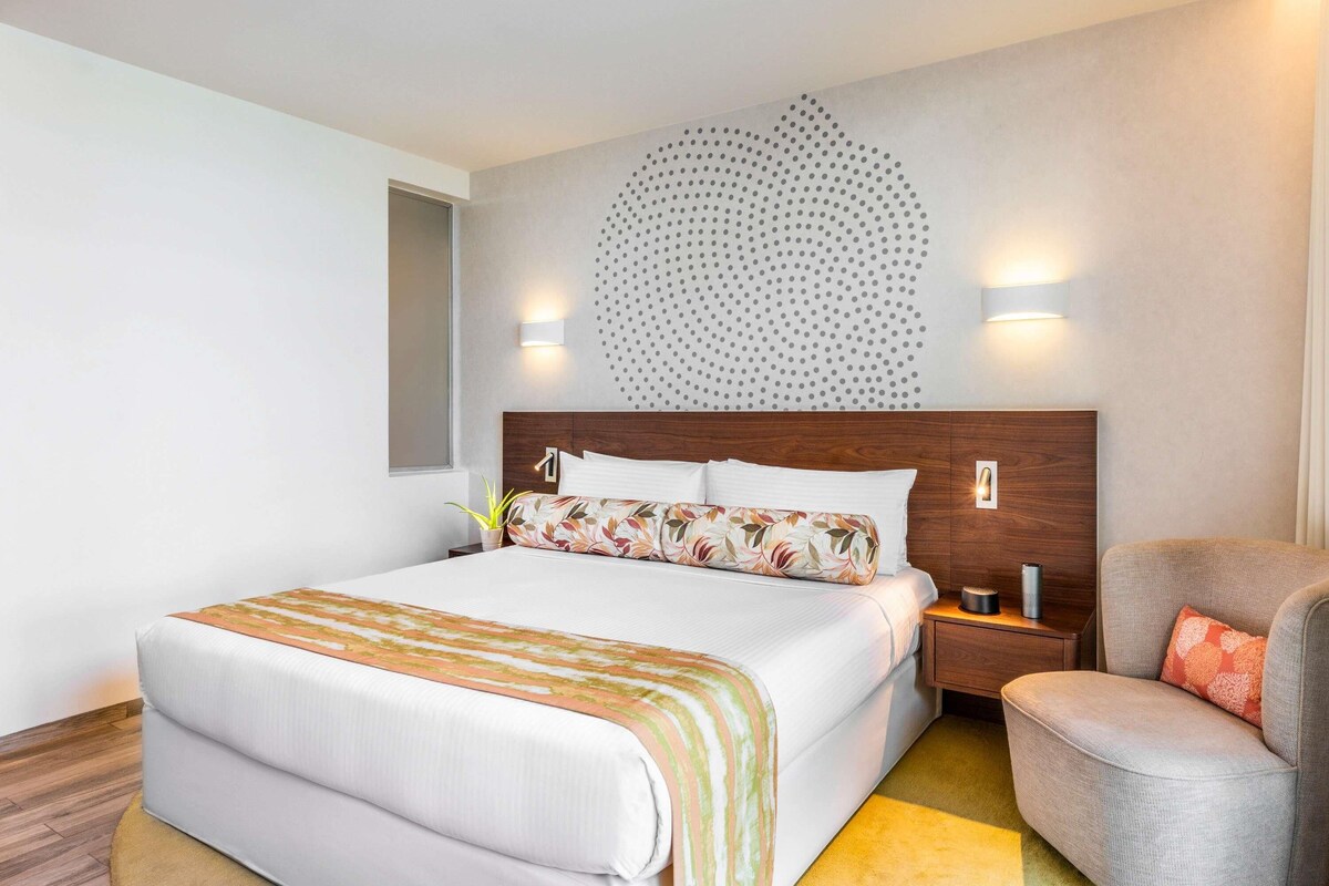A modern hotel room with a large bed, a wooden headboard, a wall-mounted light fixture, and a small side table.