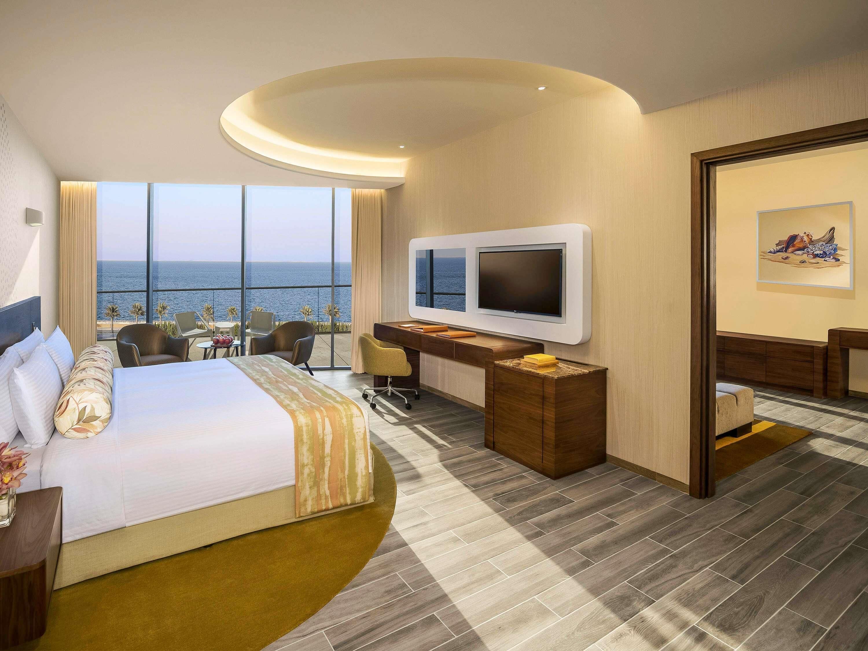 Een hotelkamer met een bed, bureau, televisie en uitzicht op zee.
