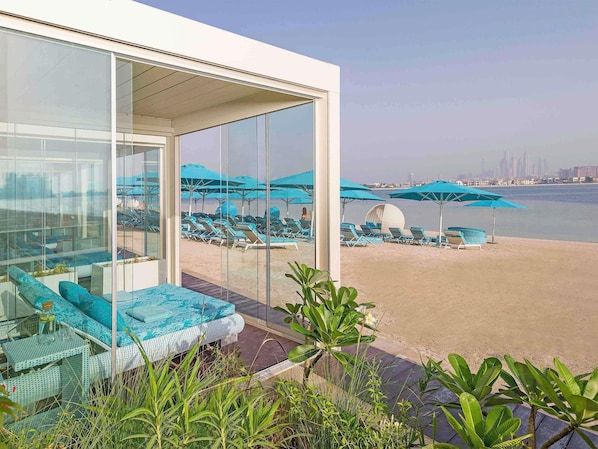 Une chambre dâhĂŽtel moderne avec un mur en verre donnant sur une plage Ă©quipĂ©e de transats et de parasols.