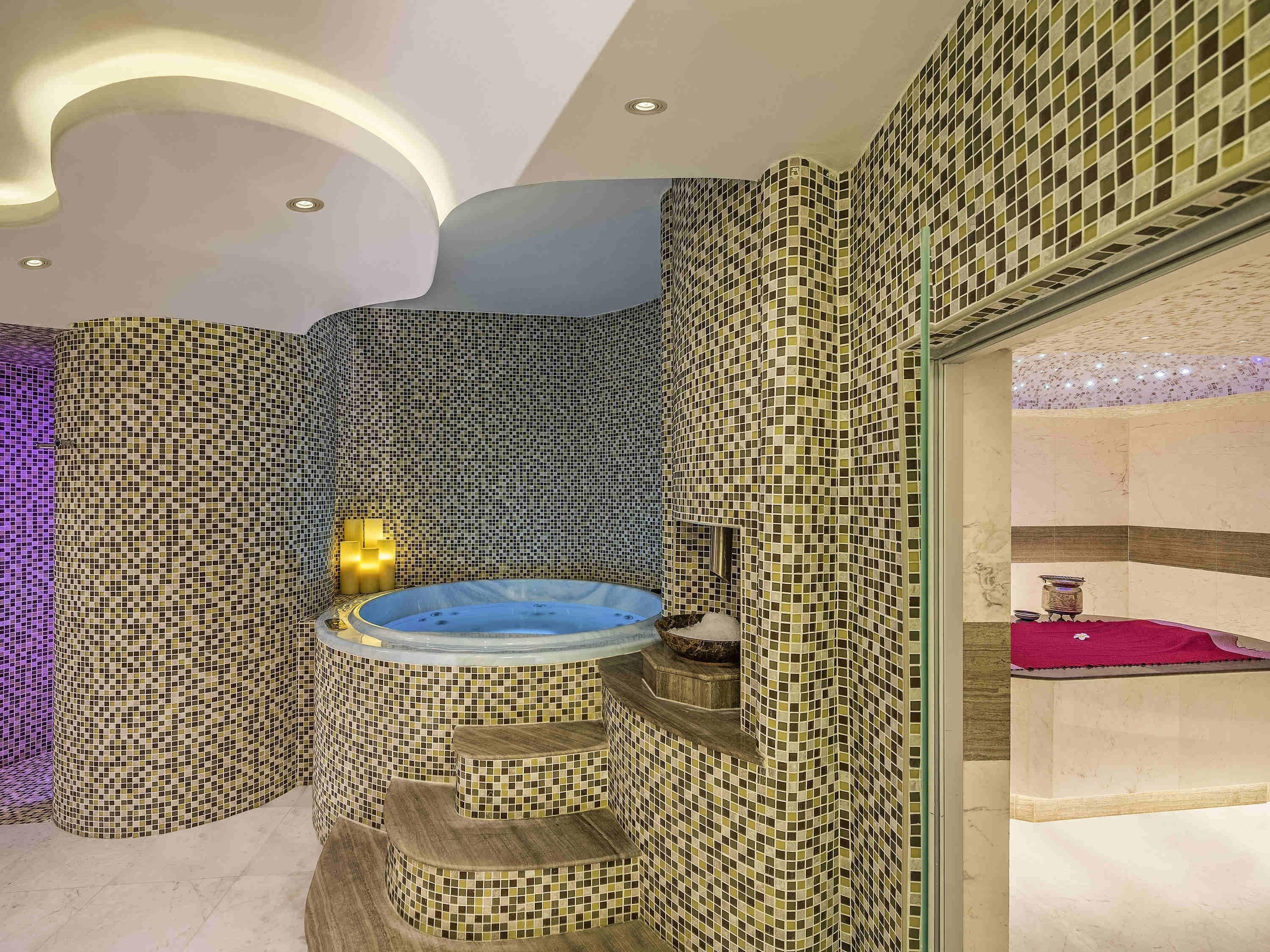 Un baño con pared de mosaicos, una bañera de hidromasaje y un lavamanos pequeño.