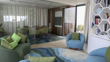 A modern hotel room with a green sofa, a blue armchair, a glass partition, a dining area with chairs, and a bookshelf.