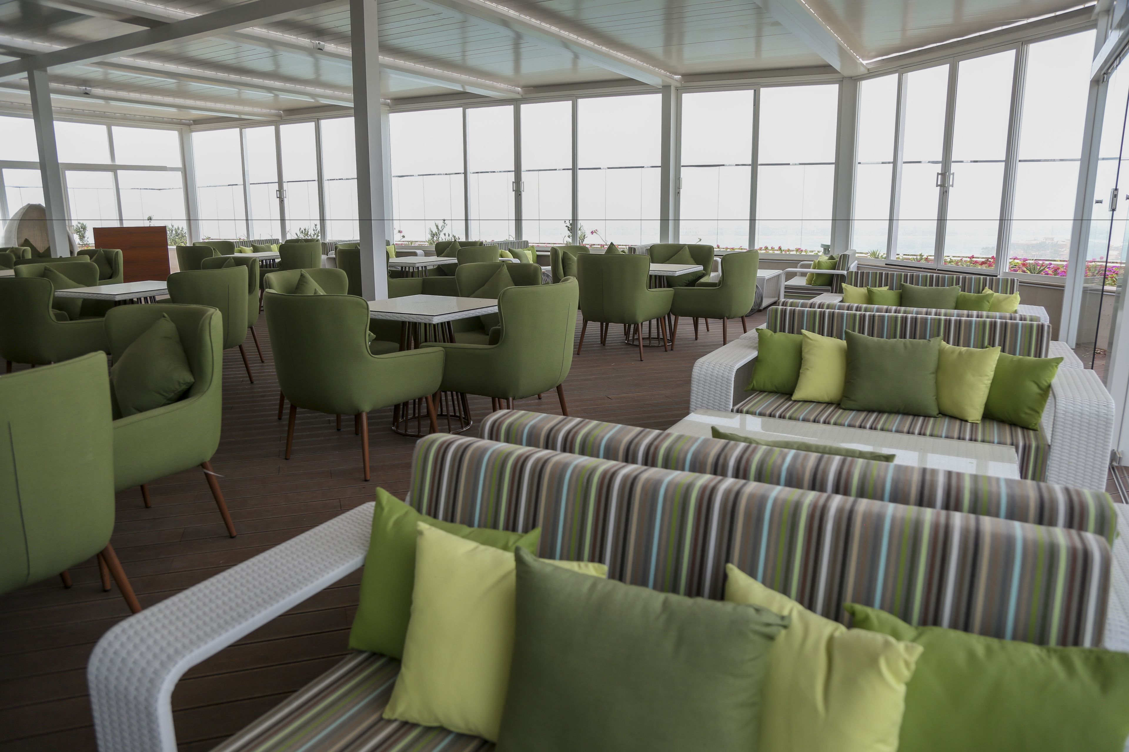 A covered outdoor seating area with green chairs and striped sofas, large windows, and a view of the city.