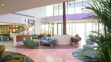 A spacious lobby with a modern design, featuring a central seating area with sofas and armchairs, a reception desk, and a large palm tree.