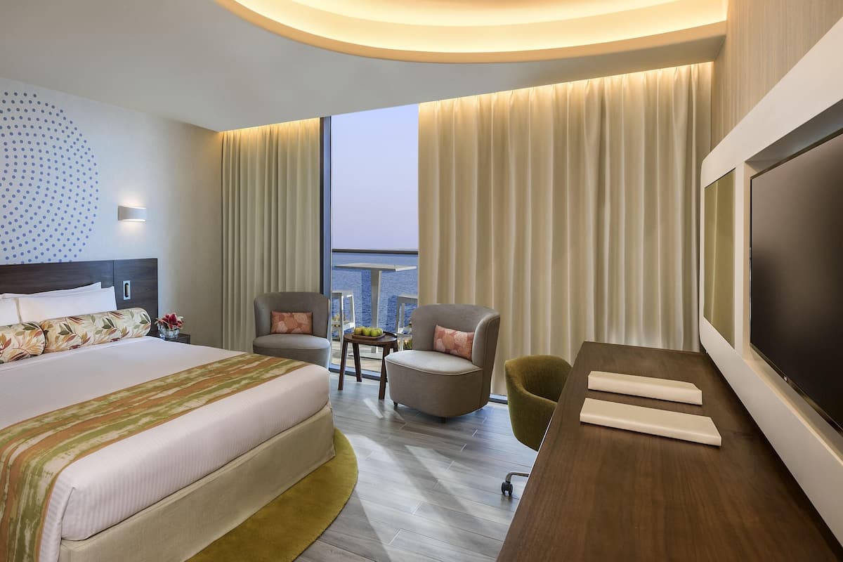 Ein modernes Hotelzimmer mit einem großen Bett, einem Schreibtisch mit Fernseher, einem Sessel und Blick auf das Wasser.