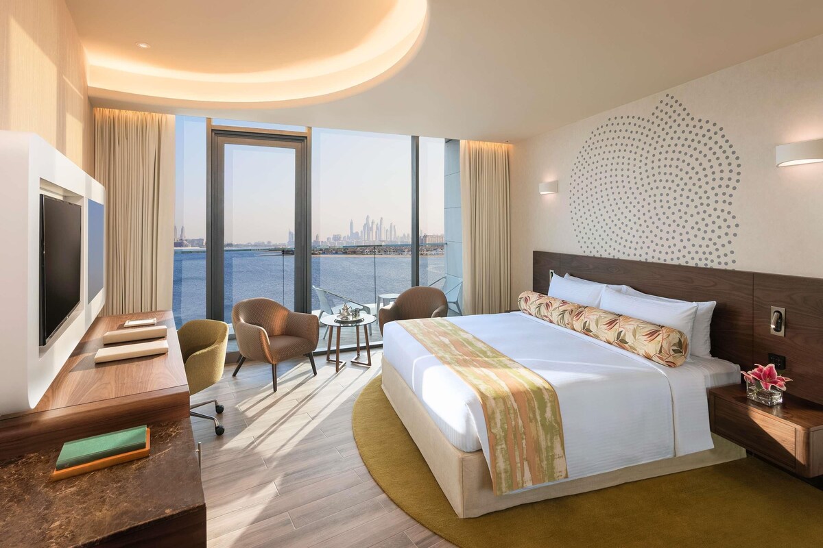 A hotel room with a large bed, a desk with a TV, a chair, a small table, and a view of the city skyline.