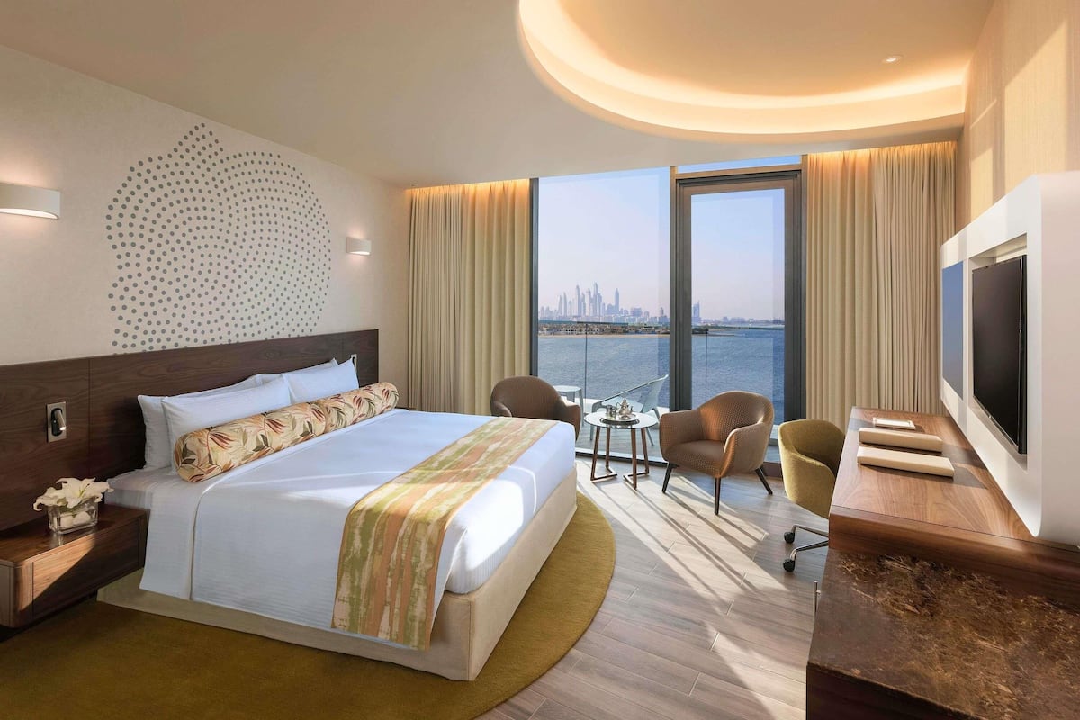 Ein modernes Hotelzimmer mit einem großen Bett, einem Schreibtisch mit Fernseher und Blick auf eine Skyline durch große Fenster.