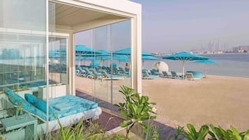 Quarto de hotel moderno com parede de vidro dando para uma praia com espreguiçadeiras e guarda-sóis.
