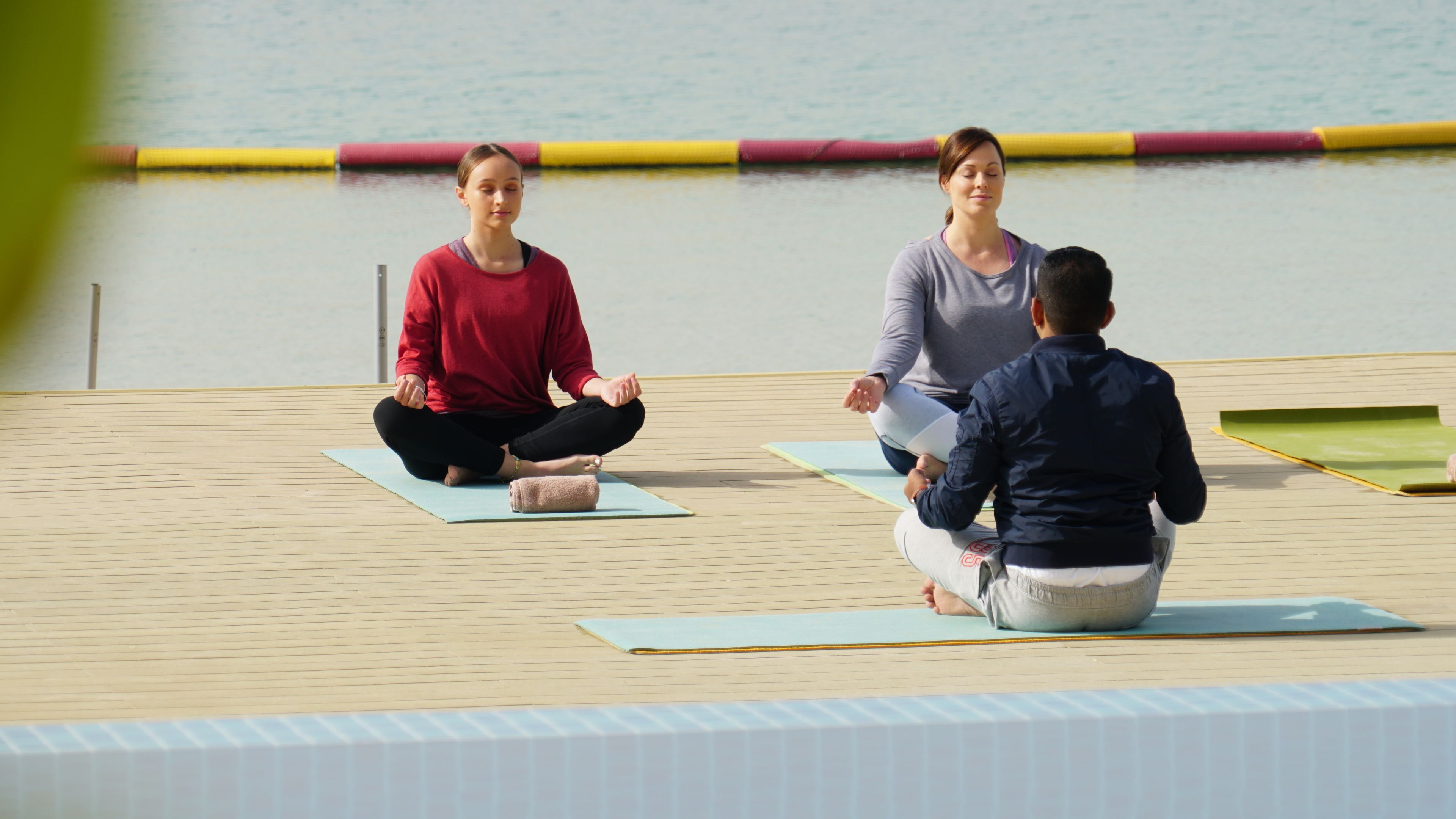 Three people sitting on yoga mats on a wooden deck by a body of water.