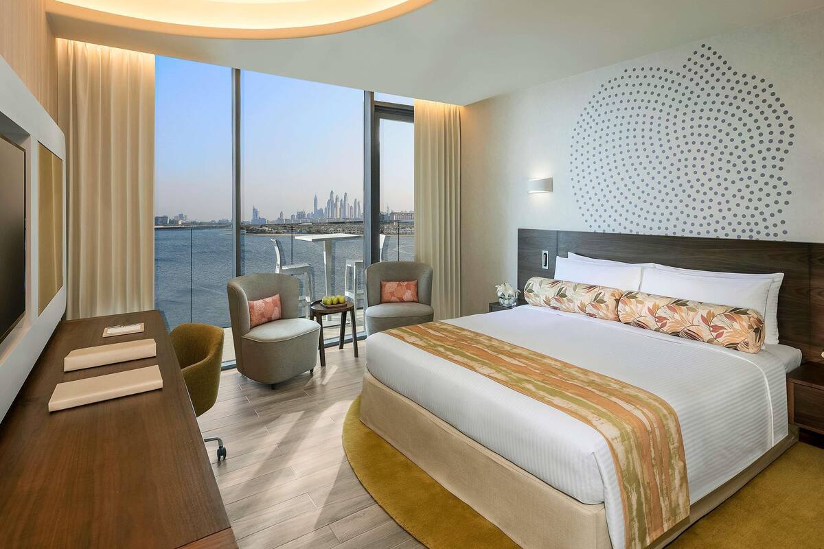 A hotel room with a large bed, a desk, two chairs, a TV, and a view of a city skyline.