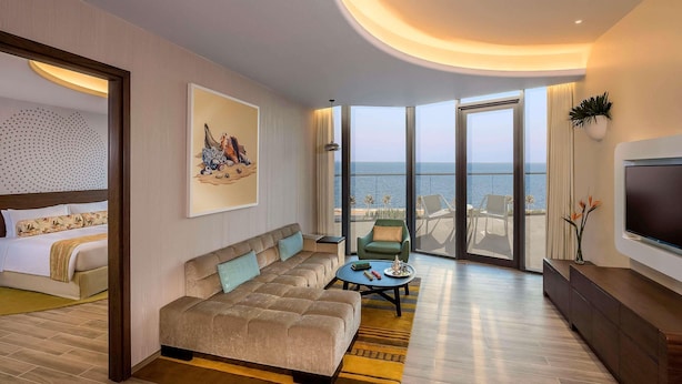 Una habitación de hotel moderna con una cama grande, un sofá, televisión y un balcón con vistas al mar.