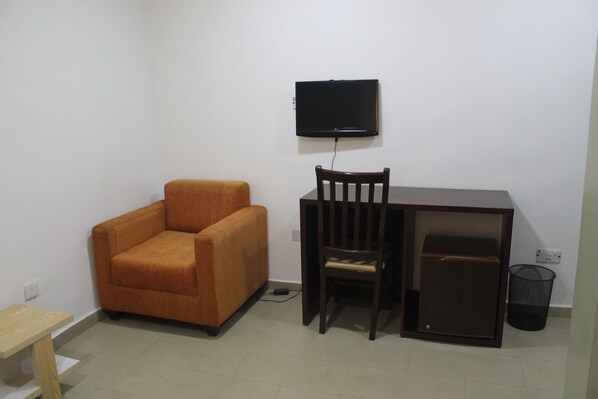 Una habitación pequeña con un escritorio de madera, una silla, un televisor fijado a la pared y un cubo de basura.