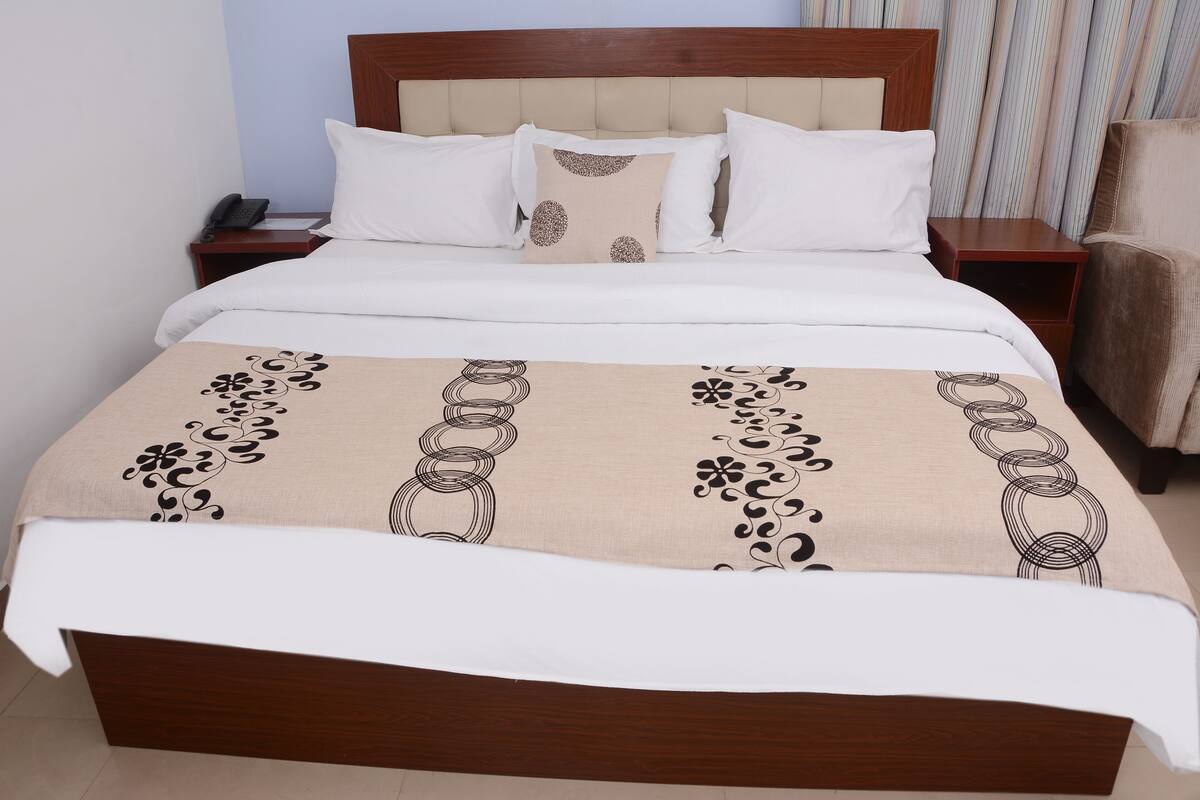 Una cama bien tendida con una manta color beige con diseños negros, cabecera de madera y almohadas blancas.