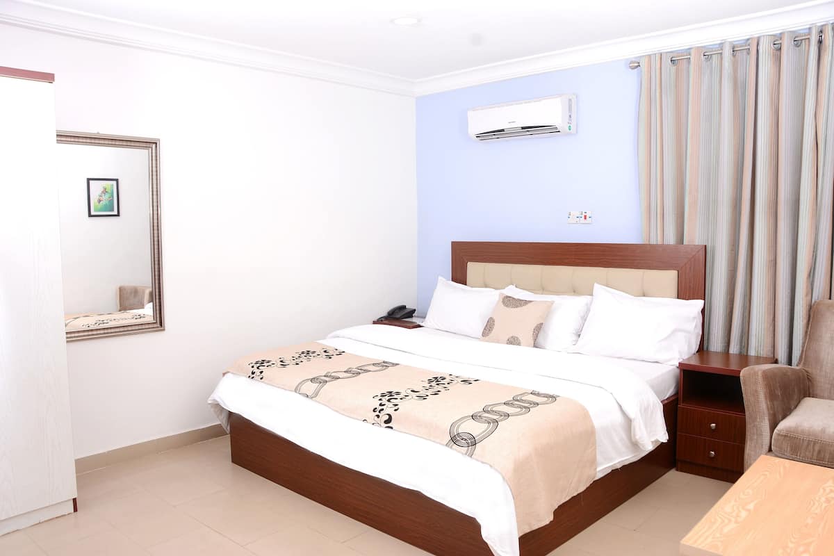 Una cama bien tendida con una colcha estampada, una mesita de noche con un teléfono y una silla en una habitación con paredes azul claro y una ventana con cortinas.