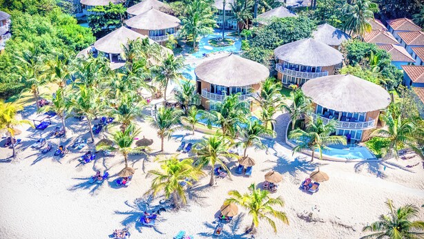Vue aérienne d’un complexe hôtelier comprenant plusieurs bungalows sur pilotis, un espace piscine central et une plage avec des chaises longues.