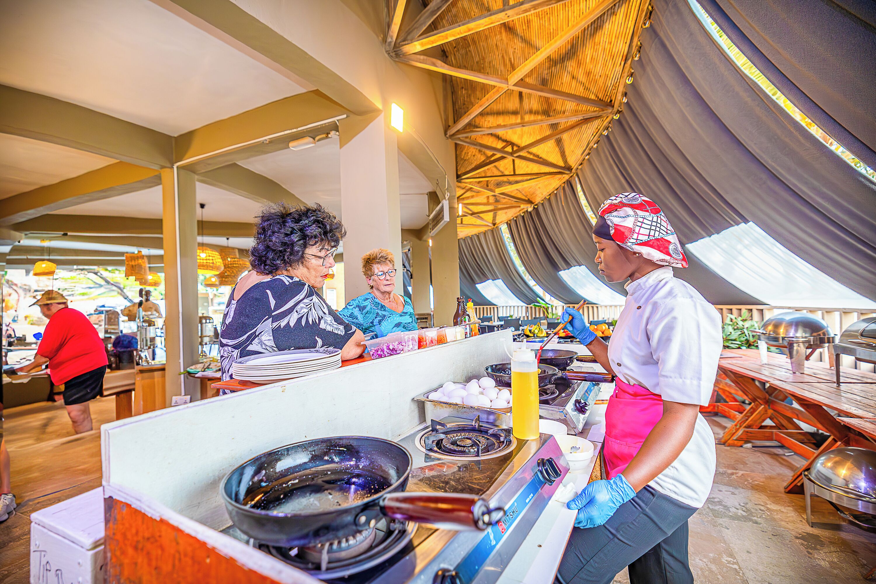 Un puesto de comida al aire libre con equipo de cocina, una persona sirviendo comida y una zona cubierta al fondo.