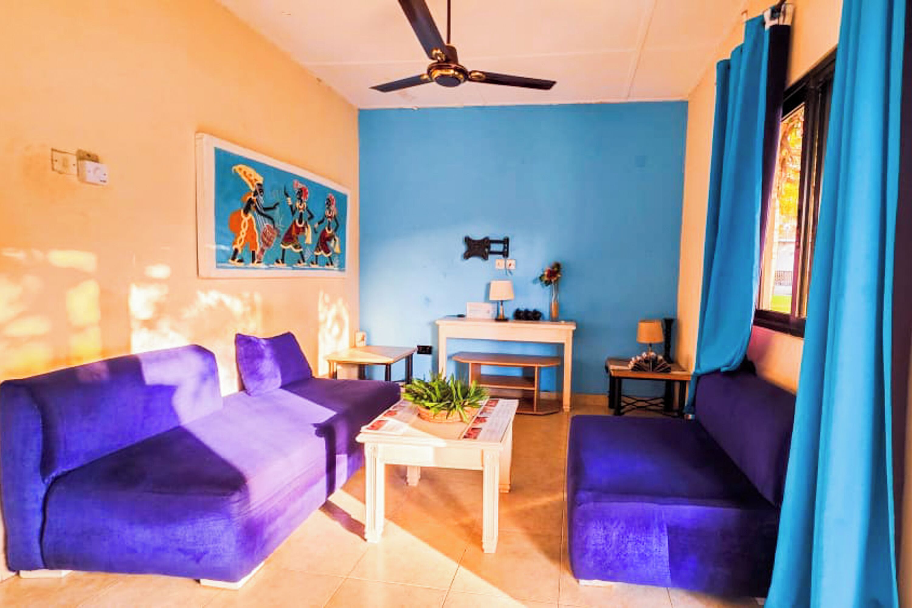 A living room with two purple sofas, a coffee table, a ceiling fan, a painting on the wall, and a window with blue curtains.