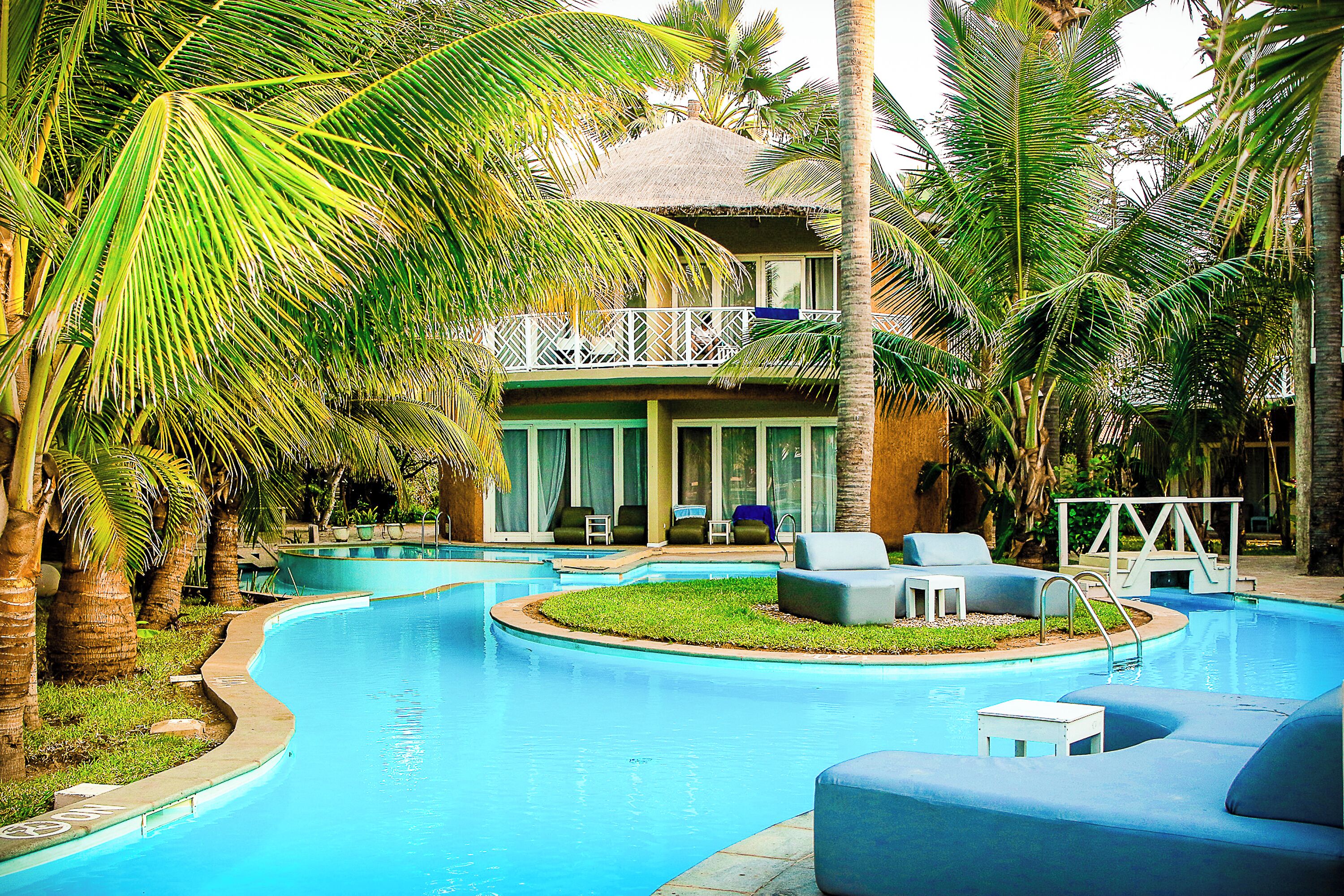 A swimming pool with lounge chairs and a thatched-roof building surrounded by palm trees.