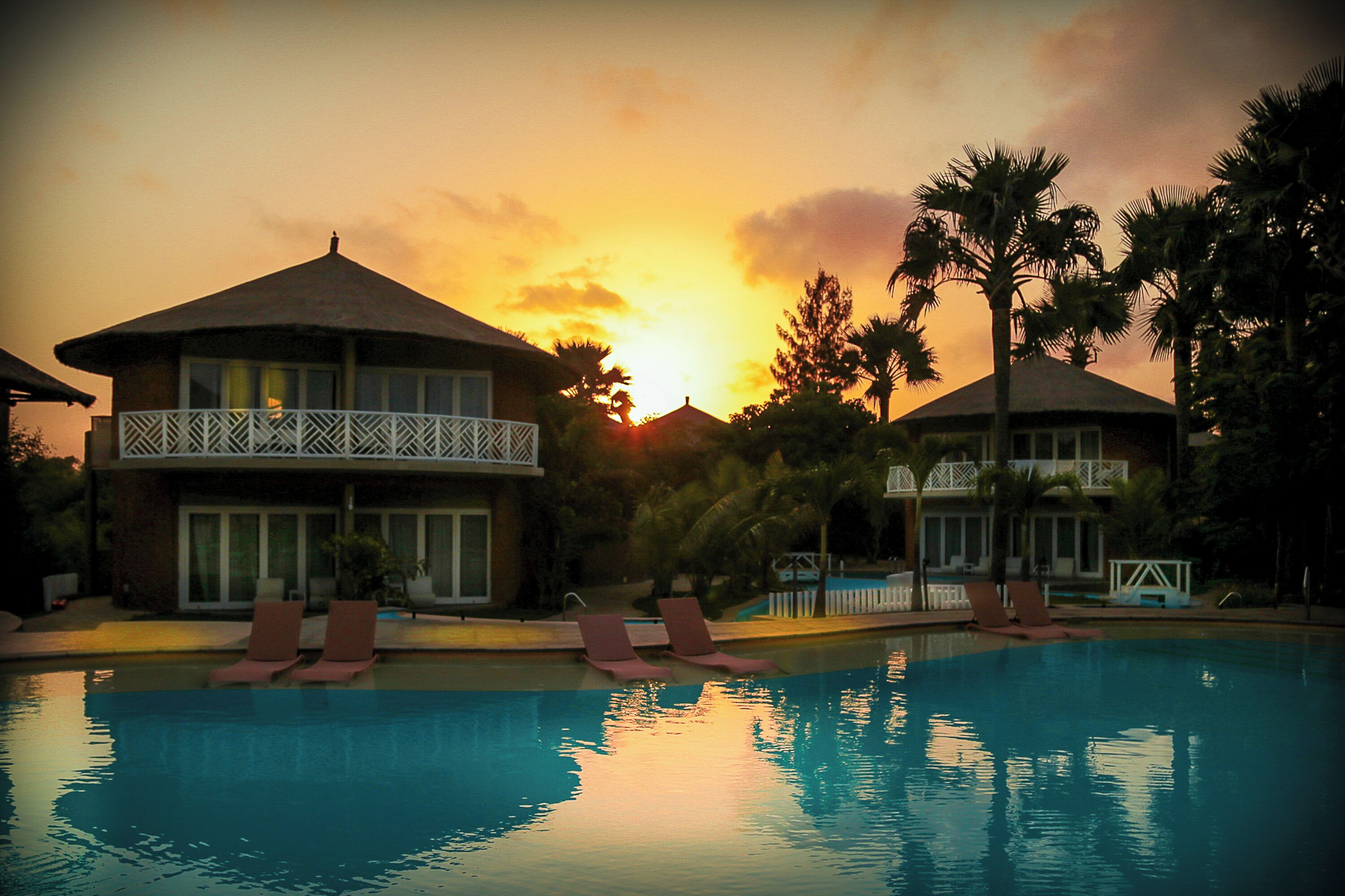 Zona de piscina con sillas de descanso y un edificio con balcón, rodeada de palmeras y un atardecer de fondo.