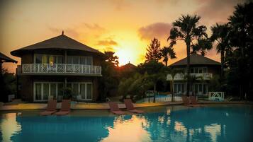 Zona de piscina con sillas de descanso y un edificio con balcón, rodeada de palmeras y un atardecer de fondo.