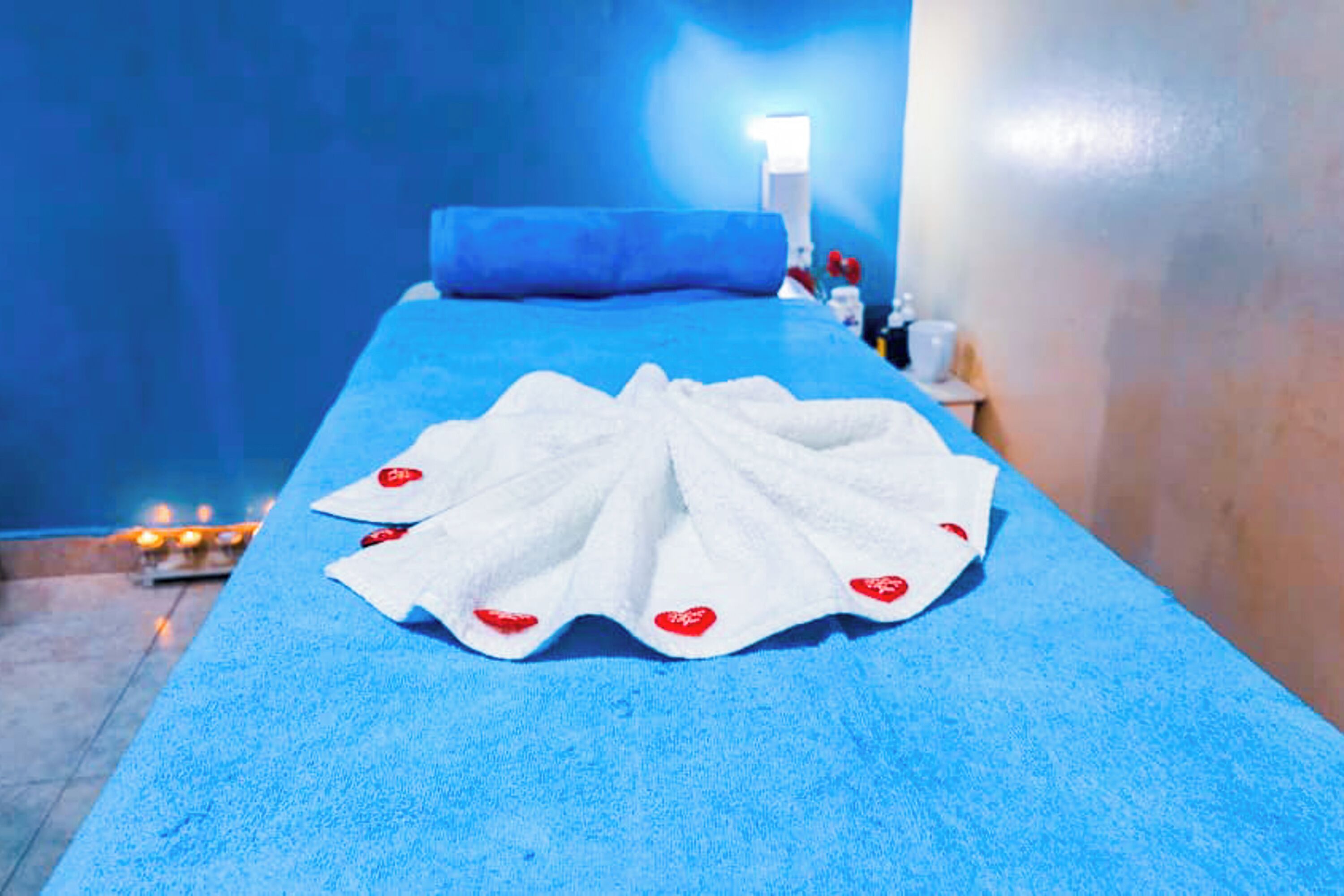 Une pièce avec une table de massage bleue, une serviette blanche ornée de motifs en forme de cœur rouge, et une petite table sur laquelle se trouvent une lampe et une bouteille.