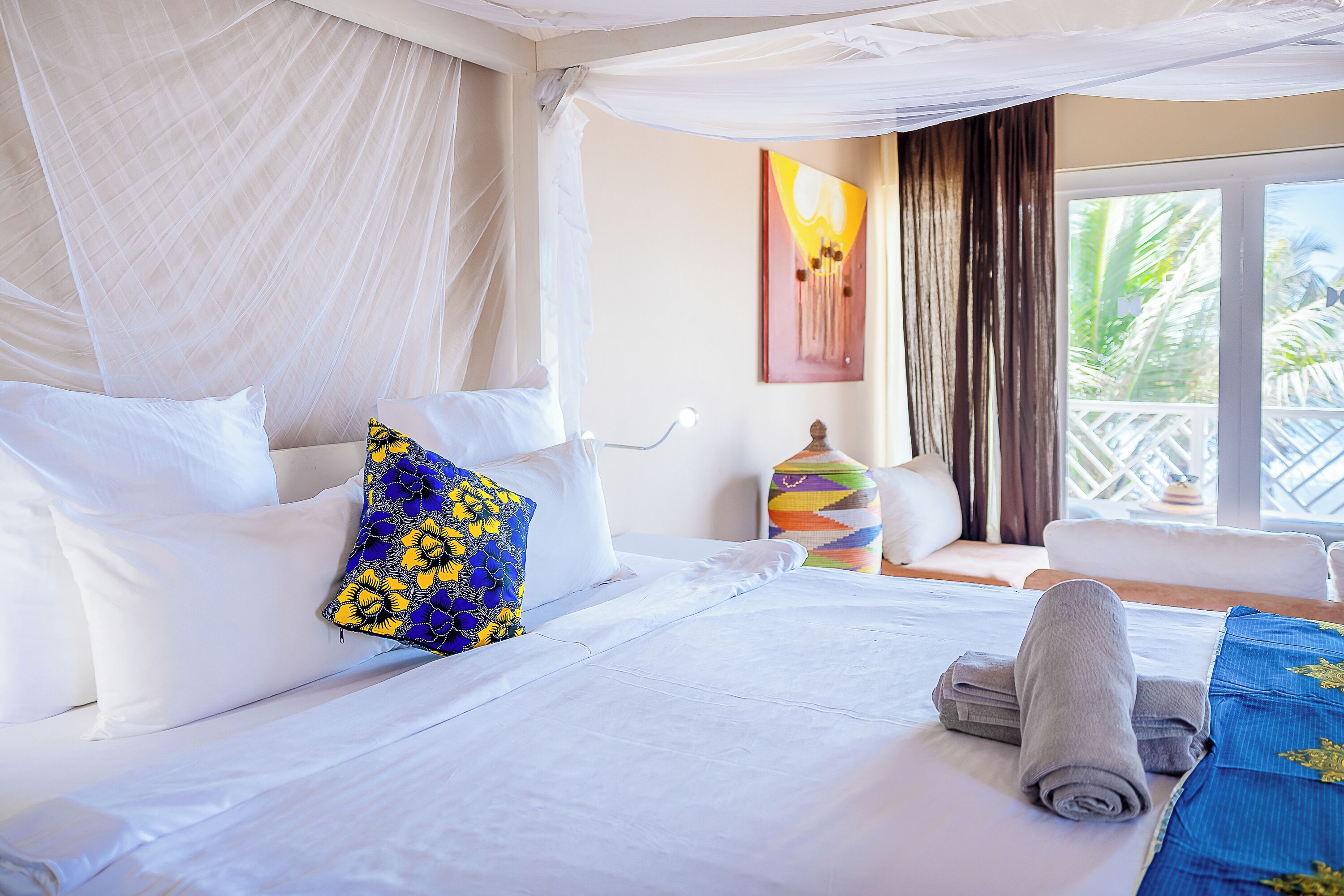 Una habitación de hotel con cama, mesita de noche, ventana con vistas a palmeras y un cuadro en la pared.