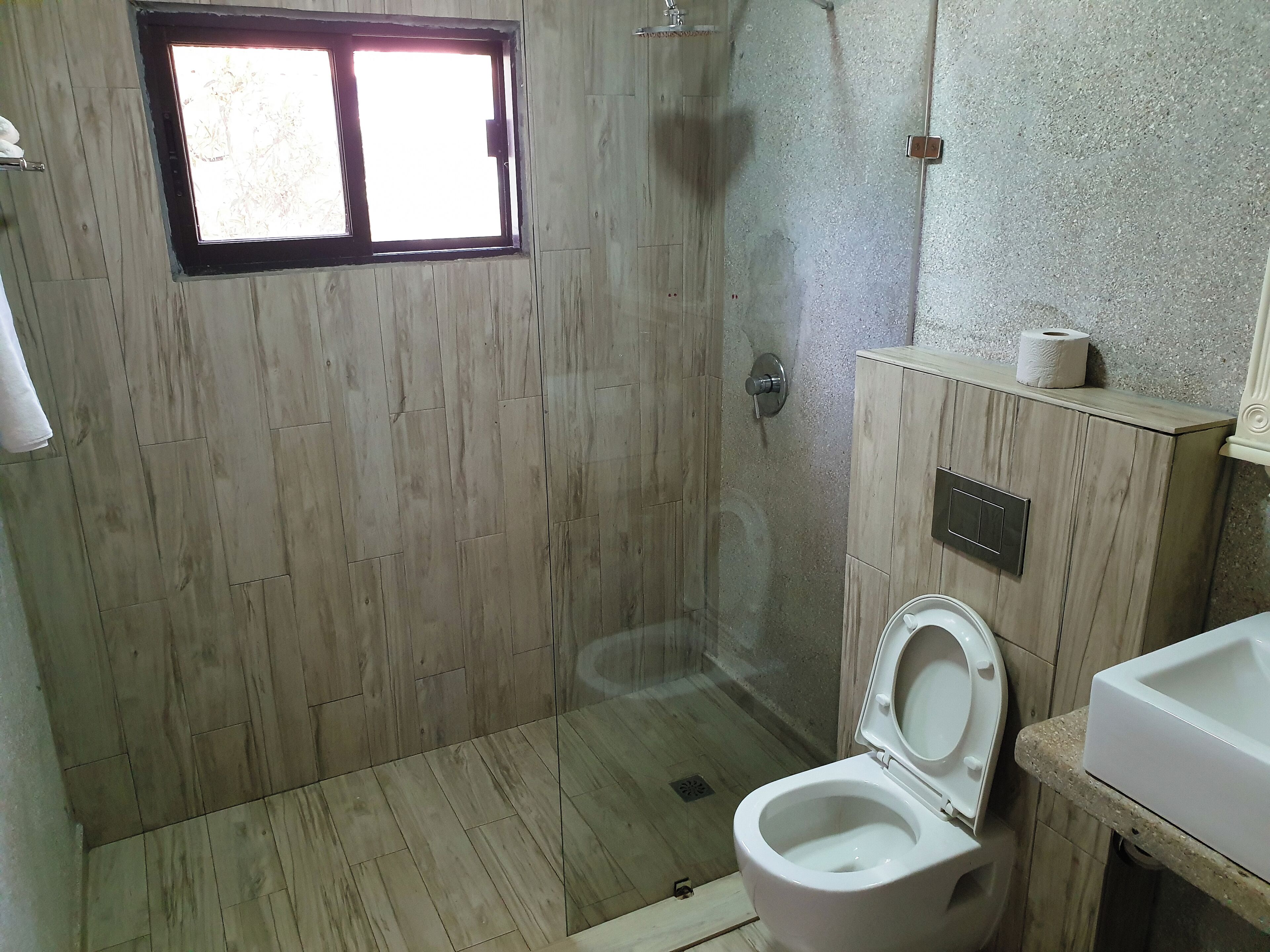 Une salle de bain avec un plancher en bois, une douche en verre, des toilettes et un meuble en bois muni d’un porte-rouleau de papier toilette.