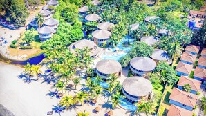 Vue aérienne d’un complexe hôtelier avec des bungalows aux toits de chaume, des palmiers et une plage.