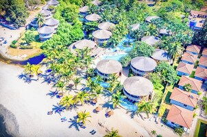 Vista aérea de un resort con bungalows de techo de paja, palmeras y una zona de playa.