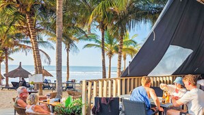Un espace de restauration aménagé sur la plage, avec des tables et des chaises, des palmiers et une tente.